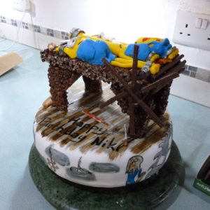 white minion cake decorated like a torture chamber. on top is a chocolate and crispie stretching rack with 3 minions stretched and various whips and implements, bottom view