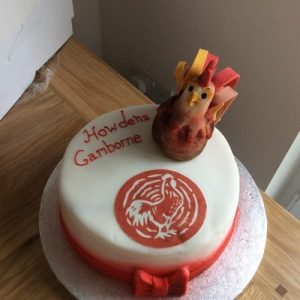 Whit cake decorated with a red bow topped with a 3D cockerel and Howdens logo